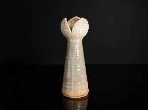 A small sculptural ceramic vase with a tall, flared, tulip-like top and a narrow tapered body, shown on a glossy black surface against a black background.