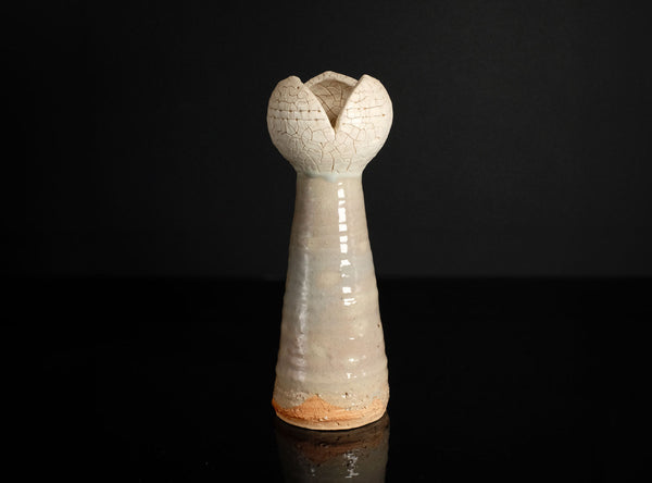 A small sculptural ceramic vase with a tall, flared, tulip-like top and a narrow tapered body, shown on a glossy black surface against a black background.