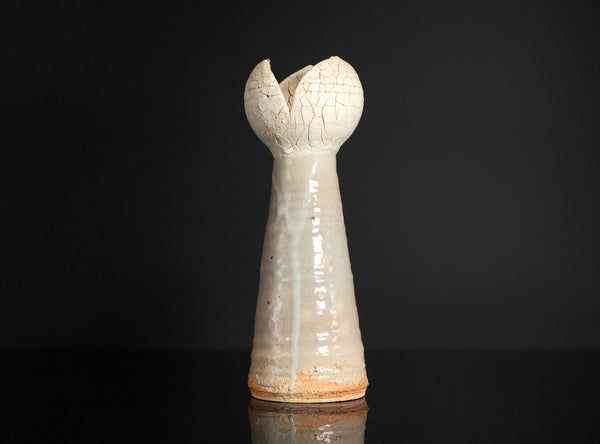 A small sculptural ceramic vase with a tall, flared, tulip-like top and a narrow tapered body, shown on a glossy black surface against a black background.