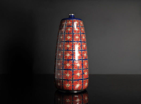 A tall, narrow ceramic vase with a glossy red and blue floral pattern, standing on a reflective black surface against a dark gray background.