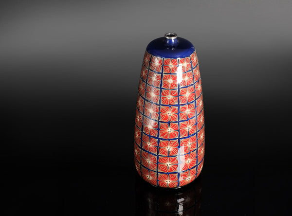 A glossy ceramic bottle with a red and blue geometric floral pattern and a dark blue lid, set against a dark gradient background.