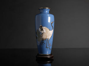A blue ceramic vase with hand-painted cranes, displayed on a dark wooden stand against a dark gray background.
