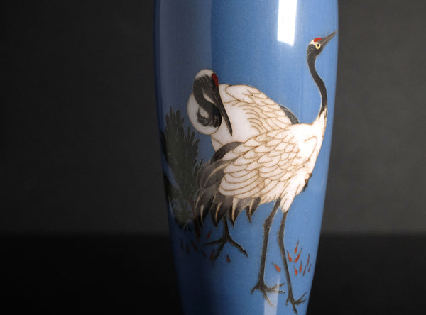A close-up of a blue ceramic vase featuring a hand-painted crane with white, black, and gray feathers, set against a dark background.