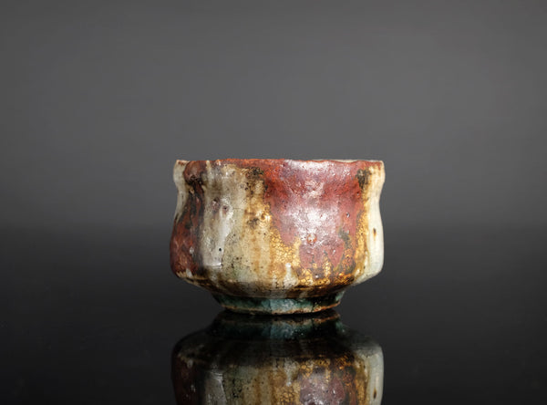 A handmade ceramic sake cup with a rustic, hand-painted glaze featuring mottled red, brown, beige, and green colors, set against a dark gray background with a reflective black surface below.