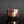 Load image into Gallery viewer, A handmade ceramic sake cup with a rustic, hand-painted glaze featuring mottled red, brown, beige, and green colors, set against a dark gray background with a reflective black surface below.
