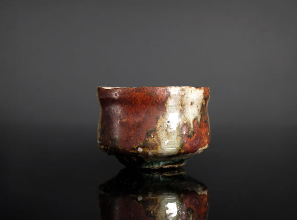 A handmade ceramic sake cup with a rustic, hand-painted glaze featuring mottled red, brown, beige, and green colors, set against a dark gray background with a reflective black surface below.