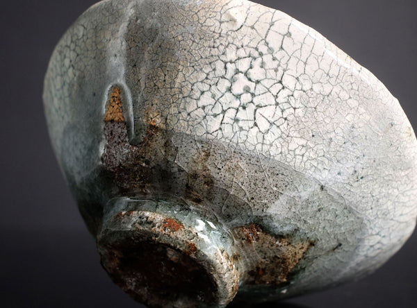 Close-up of a ceramic bowl with a glossy crackled glaze in pale gray-green and white, showing a rustic dark brown base and a small side loop, set against a dark background.