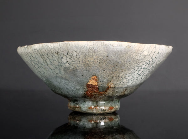 A handmade ceramic bowl with a light gray and white glaze featuring a prominent crackle pattern, resting on a dark reflective surface against a dark gray background.