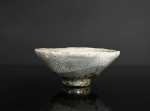 A handmade ceramic bowl with a light gray and white glaze featuring a prominent crackle pattern, resting on a dark reflective surface against a dark gray background.