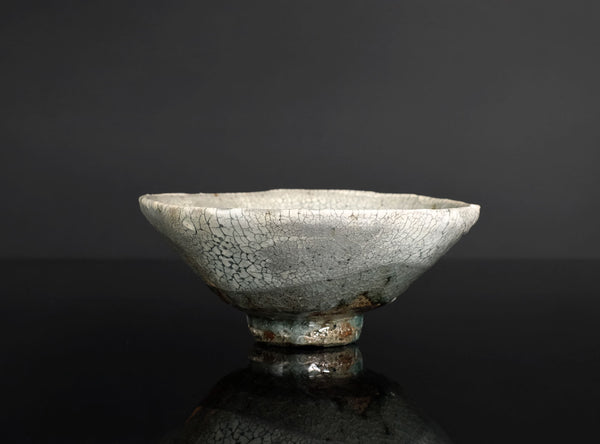 A handmade ceramic bowl with a light gray and white glaze featuring a prominent crackle pattern, resting on a dark reflective surface against a dark gray background.