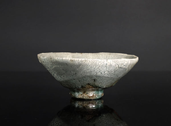 A handmade ceramic bowl with a light gray and white glaze featuring a prominent crackle pattern, resting on a dark reflective surface against a dark gray background.