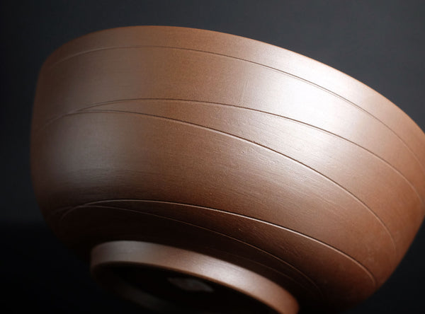 A close-up of a brown ceramic bowl with horizontal grooved lines, photographed against a dark black background.