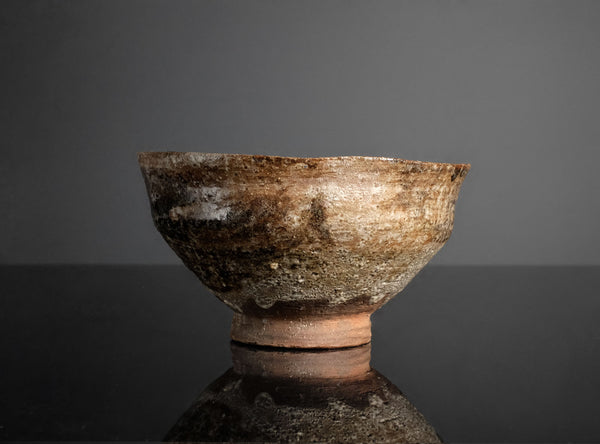 An ancient-looking rustic clay bowl with a textured, weathered surface in earthy brown, beige, and gray tones, set against a dark gray background with a reflective black surface below.