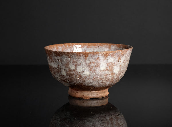A handmade ceramic bowl with a textured, mottled glaze of brown and white, standing on a small footed base against a dark reflective background.