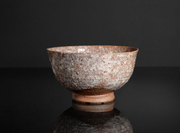 A rustic handmade ceramic bowl with a textured, speckled glaze in earthy brown, beige, and gray tones, photographed on a dark reflective surface against a black background.