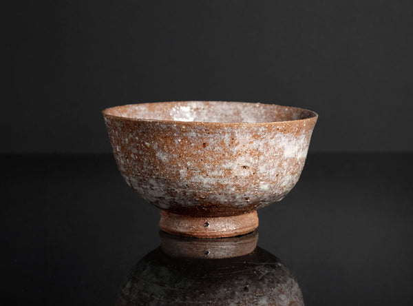 A handmade ceramic bowl with a textured, speckled brown and white glaze, standing on a small matching base against a dark reflective background.