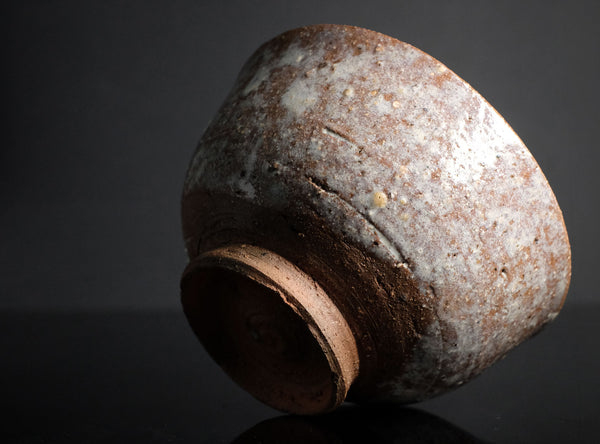 A rustic handmade ceramic bowl with a textured, speckled glaze in earthy brown, beige, and gray tones, photographed on a dark reflective surface against a black background.