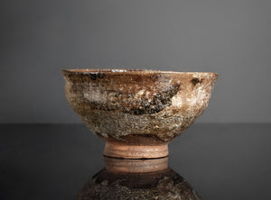 An antique ceramic bowl with a rustic, weathered finish, featuring a brown and beige textured glaze with dark patches, set against a dark gray background on a reflective black surface.