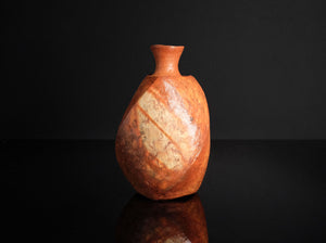 A handmade ceramic bottle with a unique organic, marbled texture in earthy terracotta orange and beige tones, set against a solid black background with a reflective surface below.
