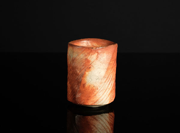 A small cylindrical cup made of marbled orange, peach, and cream ceramic, standing on a reflective black surface against a black background.