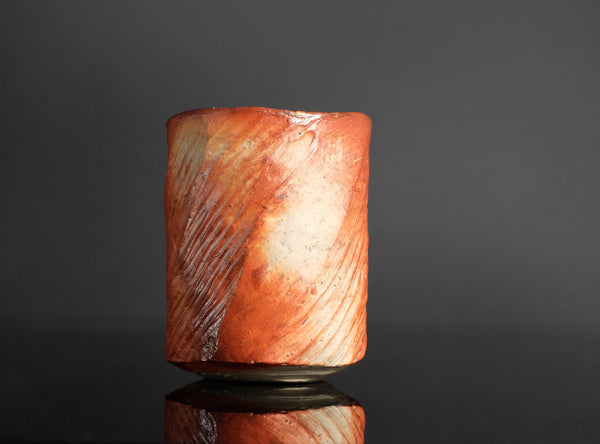 A small cylindrical cup made of marbled orange, peach, and cream ceramic, standing on a reflective black surface against a black background.