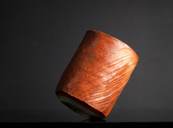 A small cylindrical cup made of marbled orange, peach, and cream ceramic, standing on a reflective black surface against a black background.