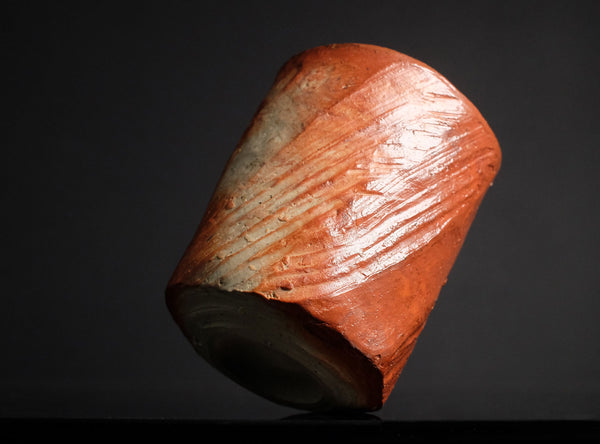 A small cylindrical cup made of marbled orange, peach, and cream ceramic, standing on a reflective black surface against a black background.
