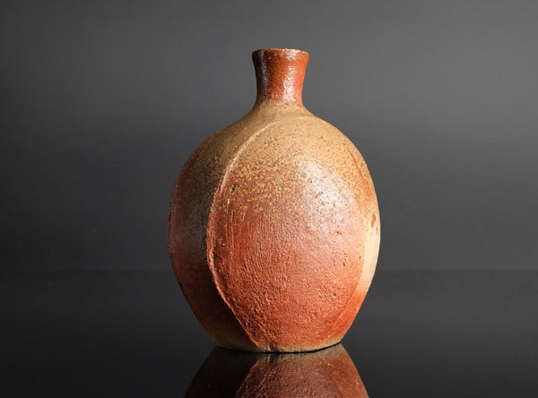 A handmade ceramic bottle with a rounded body and short neck, featuring a textured glaze that transitions from reddish-brown to light beige, set against a dark gray background.