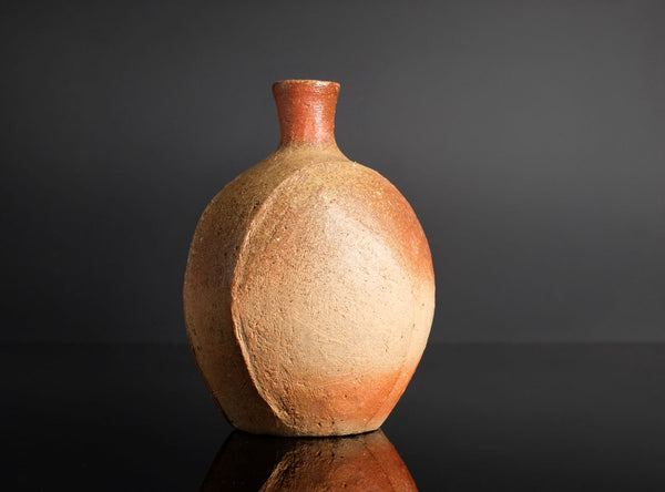A handmade ceramic bottle with a rounded body and a short, narrow neck, featuring a textured earthy brown and orange glaze, set against a dark gray background with a reflective black surface below.