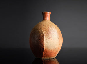 A handmade ceramic bottle with a rounded body and a short, narrow neck, featuring a textured earthy glaze with a gradient from reddish-brown at the top to beige at the bottom, set against a dark gray background.