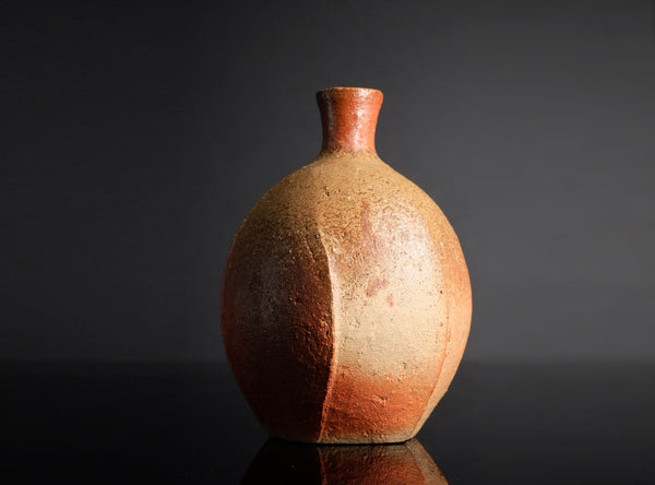 A handmade ceramic bottle with a rounded body and a short, narrow neck, featuring a textured earthy glaze with a gradient from reddish-brown at the top to beige at the bottom, set against a dark gray background.