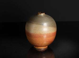 A round ceramic vase with a narrow neck, featuring a glossy glaze that transitions from beige at the top to a warm terracotta orange at the bottom, set against a dark reflective background.