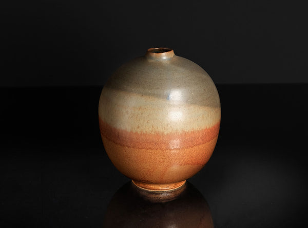 A round ceramic vase with a narrow neck, featuring a glossy glaze that transitions from beige at the top to a warm terracotta orange at the bottom, set against a dark reflective background.