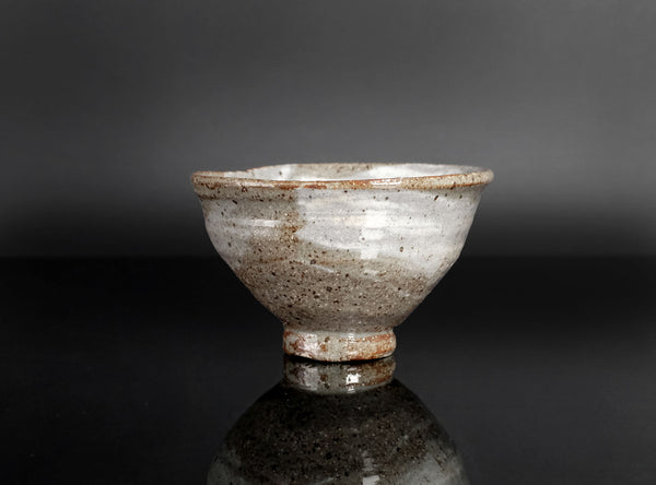 A handmade ceramic bowl with a textured, speckled glaze in shades of gray, brown, and white, standing on a dark reflective surface against a dark gray background.