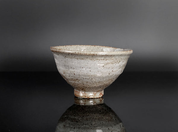 A handmade ceramic tea bowl with a textured, speckled glaze in light gray and brown tones, displayed on a reflective black surface against a dark gray background.