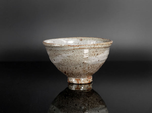 A handmade ceramic bowl with a textured, speckled glaze in earthy beige, brown, and gray tones, set against a dark gray gradient background with a reflective surface below.
