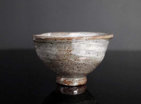 A handmade ceramic tea bowl with a textured, speckled glaze in off-white and brown tones, set against a dark gray background.