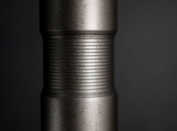 Close-up of a dark metallic cylindrical flower vase standing upright on a reflective black surface against a dark gray gradient background.