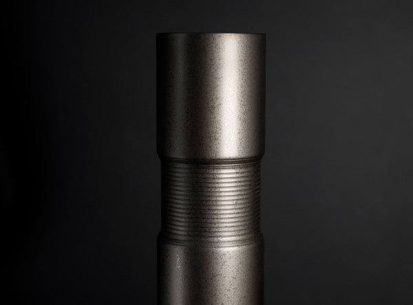 A close-up studio shot of a dark metallic cylindrical flower vase standing upright on a reflective black surface against a dark gray gradient background.
