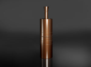 A metallic copper-colored cylindrical container or grinder with a small top tube, standing upright on a reflective dark surface against a dark gray background.