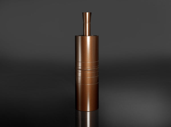 A metallic copper-colored cylindrical container or grinder with a small top tube, standing upright on a reflective dark surface against a dark gray background.