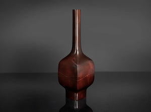 A glossy dark reddish-brown ceramic vase with a narrow neck and bulbous body, standing on a reflective black surface against a dark gray background.