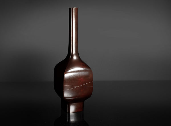A glossy dark reddish-brown ceramic vase with a narrow neck and bulbous body, standing on a reflective black surface against a dark gray background.