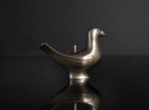 A metallic bird-shaped object with a smooth matte finish, featuring a beak, body, and a curved tail opening, photographed on a glossy black reflective surface against a black background.