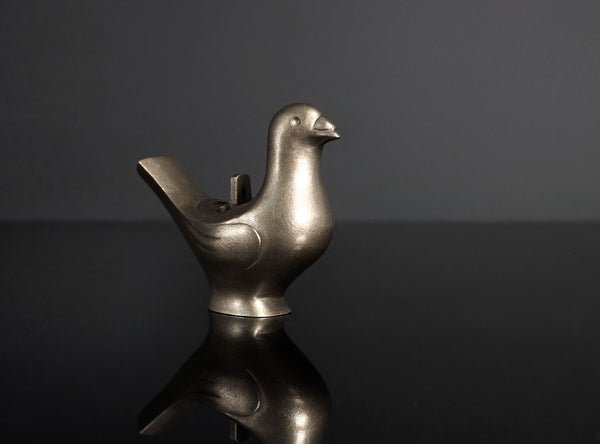 A metallic bird-shaped object with a smooth matte finish, featuring a beak, body, and a curved tail opening, photographed on a glossy black reflective surface against a black background.
