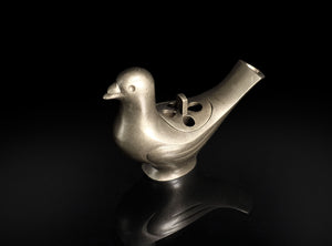 A metallic bird-shaped object with a smooth matte finish, featuring a beak, body, and a curved tail opening, photographed on a glossy black reflective surface against a black background.