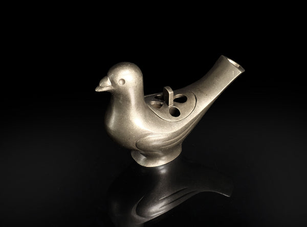 A metallic bird-shaped object with a smooth matte finish, featuring a beak, body, and a curved tail opening, photographed on a glossy black reflective surface against a black background.