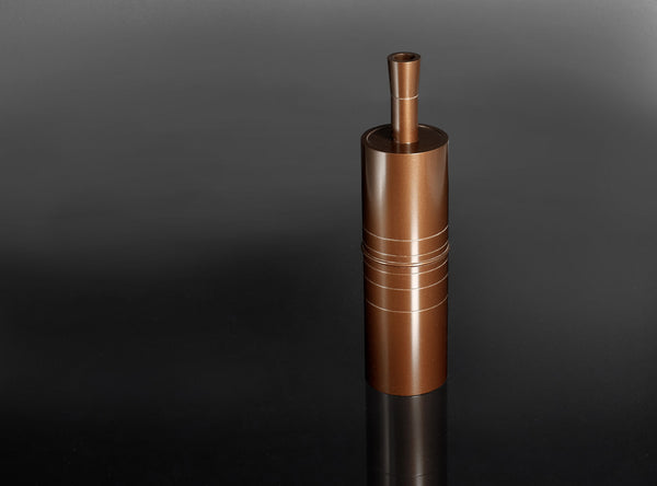 A metallic copper-colored cylindrical container or grinder with horizontal grooved details, standing upright on a reflective black surface against a dark gray gradient background.