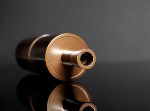 A close-up of a metallic bronze cylindrical object with a circular base and a protruding cylindrical opening, photographed on a dark reflective surface against a dark gray background.