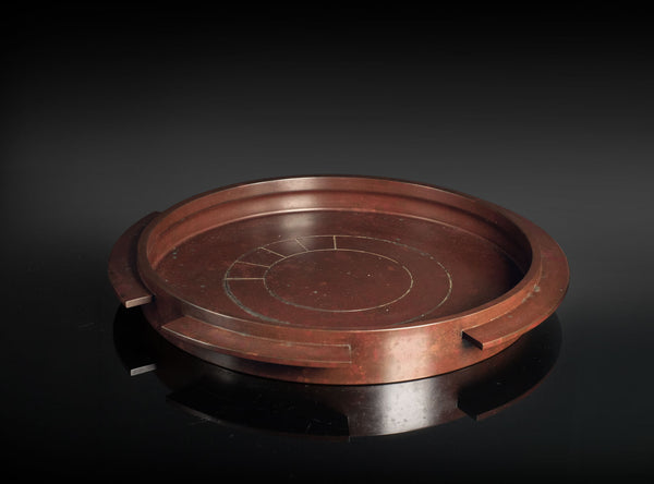 A round reddish-brown cast bronze tray with a segmented rim and engraved concentric circle details, displayed on a dark reflective surface.